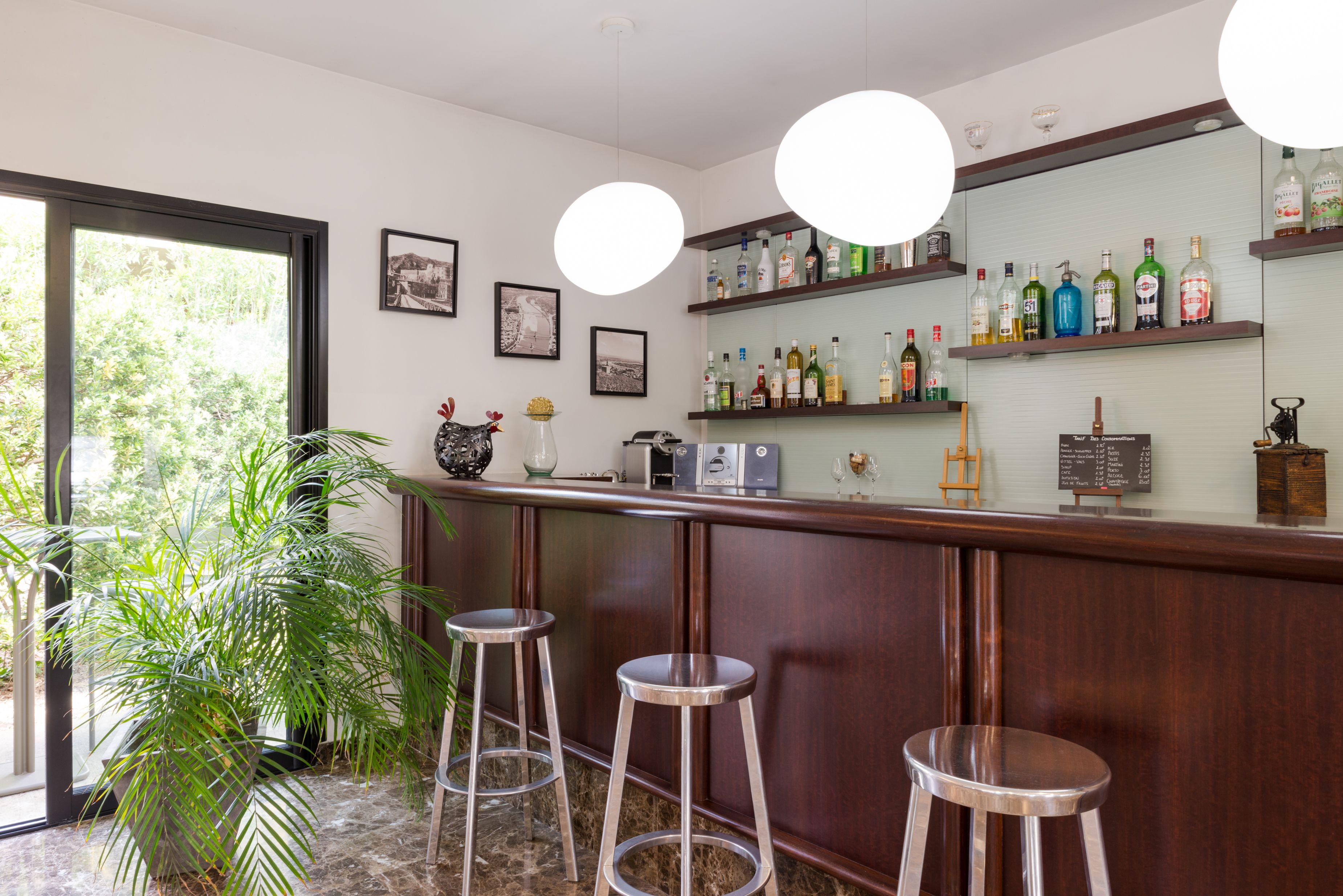 A bar area with a wooden counter, three metal stools, a potted plant, and shelves with liquor bottles.