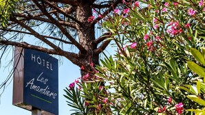 Une enseigne indiquant l’hôtel Les Amandiers, entourée d’une végétation luxuriante et de fleurs roses.