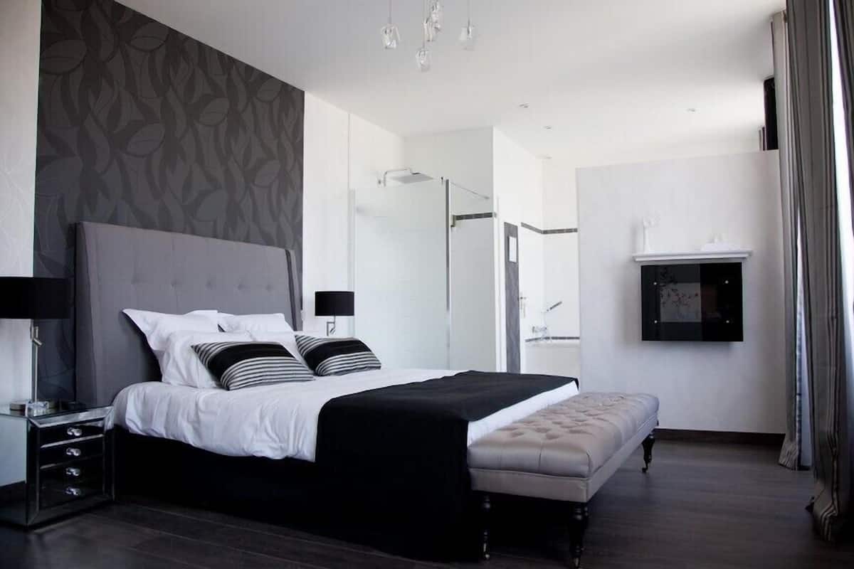 Une chambre moderne avec un grand lit, des tables de chevet, un banc, un dressing et une salle de bain avec douche et miroir.