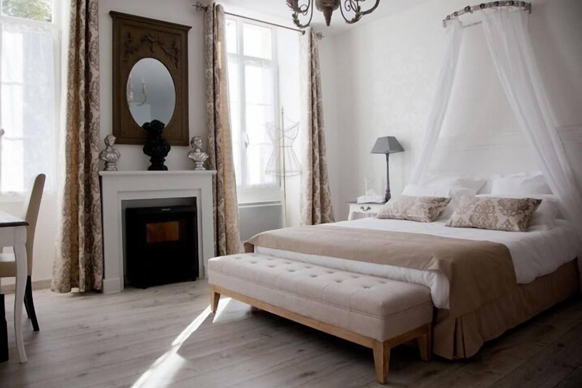 Une chambre à coucher avec un grand lit, une table de chevet, une chaise, une cheminée et une fenêtre avec des rideaux.