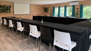 A conference room with a long table, white and black chairs, a projector screen, and windows with curtains.