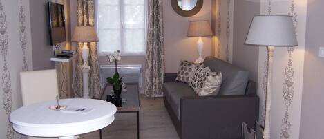A living room with a grey sofa, a white round table, and two lamps. There is a window with curtains, a mirror on the wall, and a small plant on a side table.