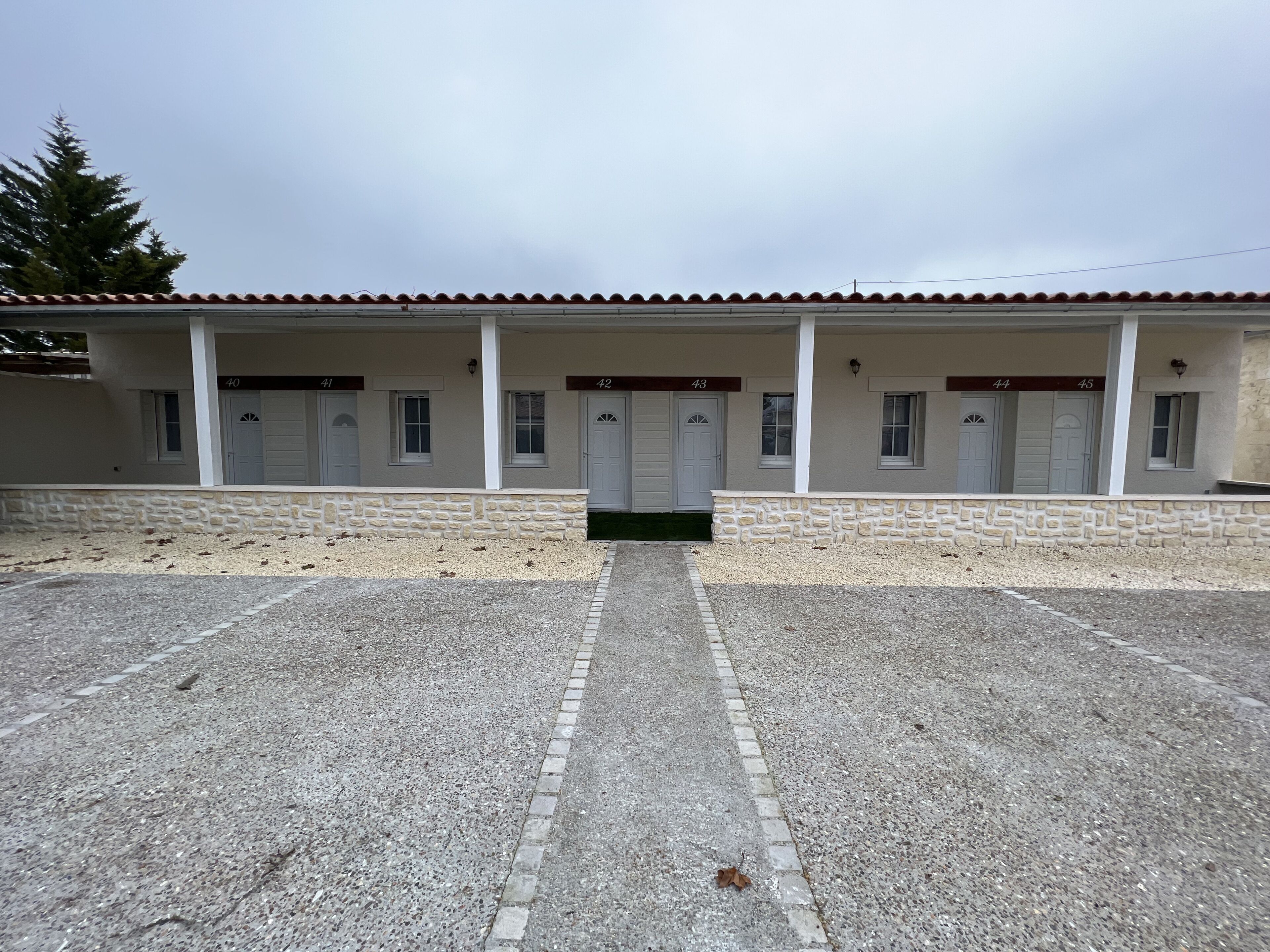 A row of white buildings with numbered doors, a stone wall, and a gravel driveway.