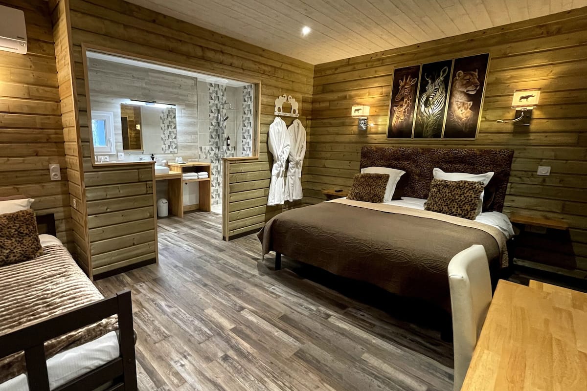 Une chambre à coucher de style cabane en bois, avec un lit, une chaise, une petite table et une salle de bain visible à travers une porte ouverte.
