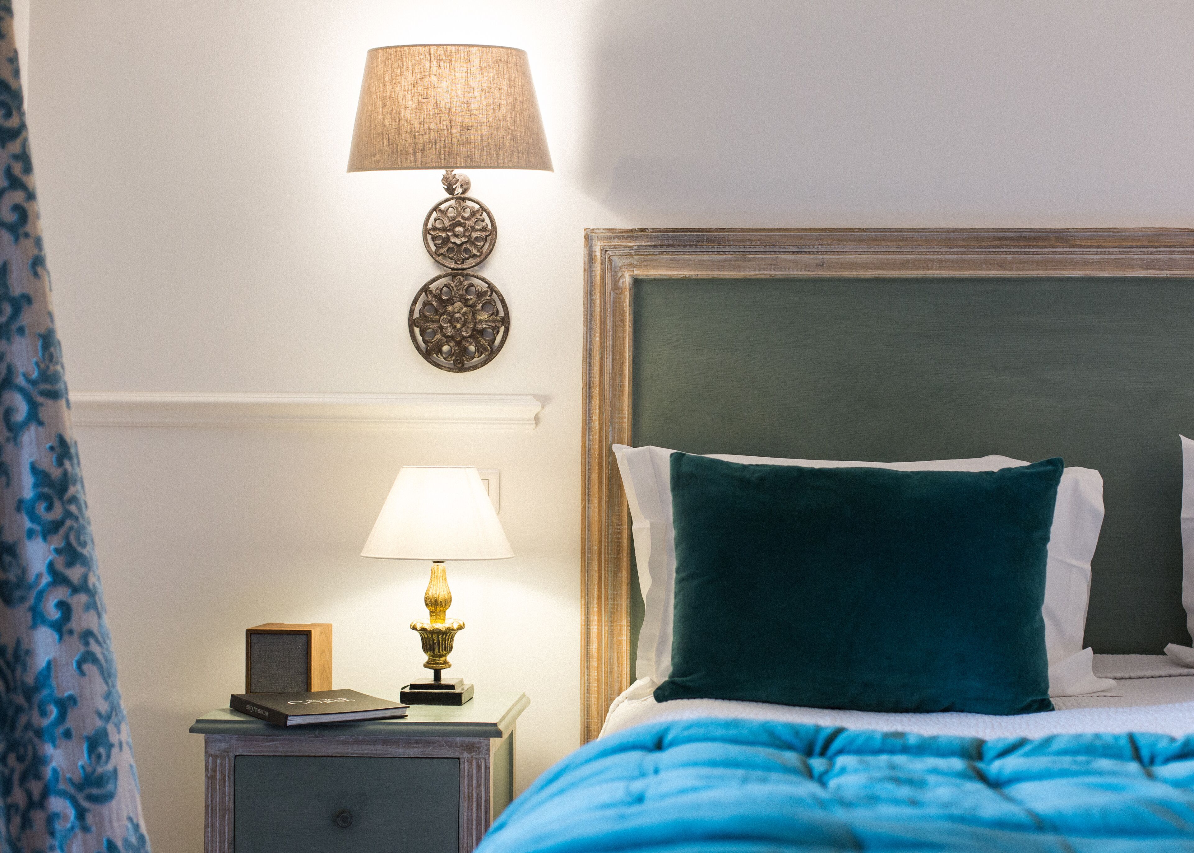 Un lit bien fait, agrémenté d’un coussin turquoise, d’une lampe de chevet et d’une lampe fixée au mur.