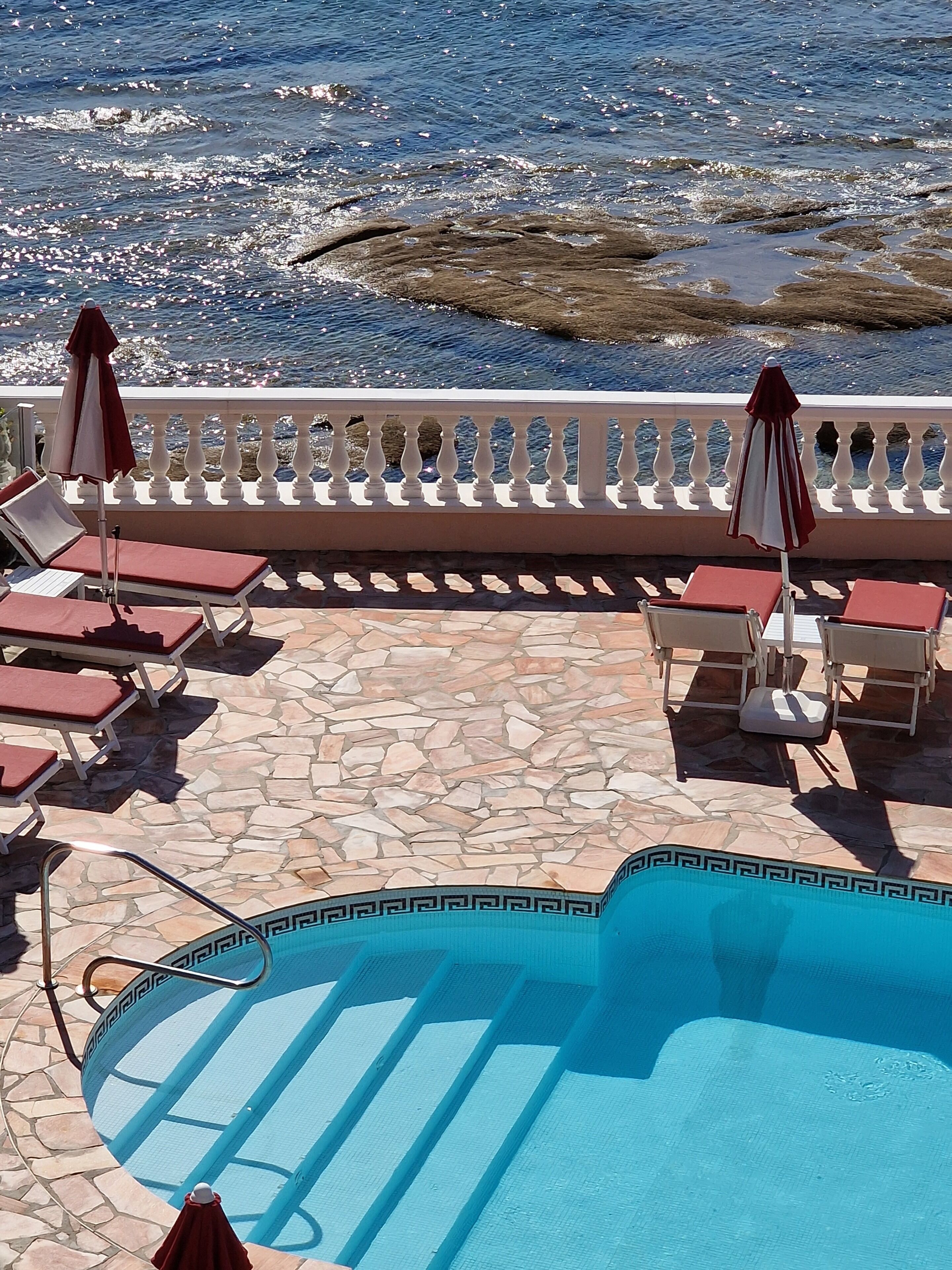 A poolside area with lounge chairs and umbrellas, a stone-tiled deck, and a view of the sea.