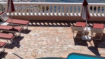 A poolside area with lounge chairs and umbrellas, a stone-tiled deck, and a view of the sea.