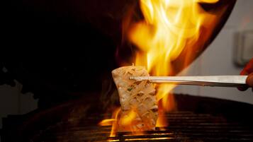 Un aliment grillé, maintenu à l’aide de pinces au-dessus d’un gril où l’on voit des flammes.