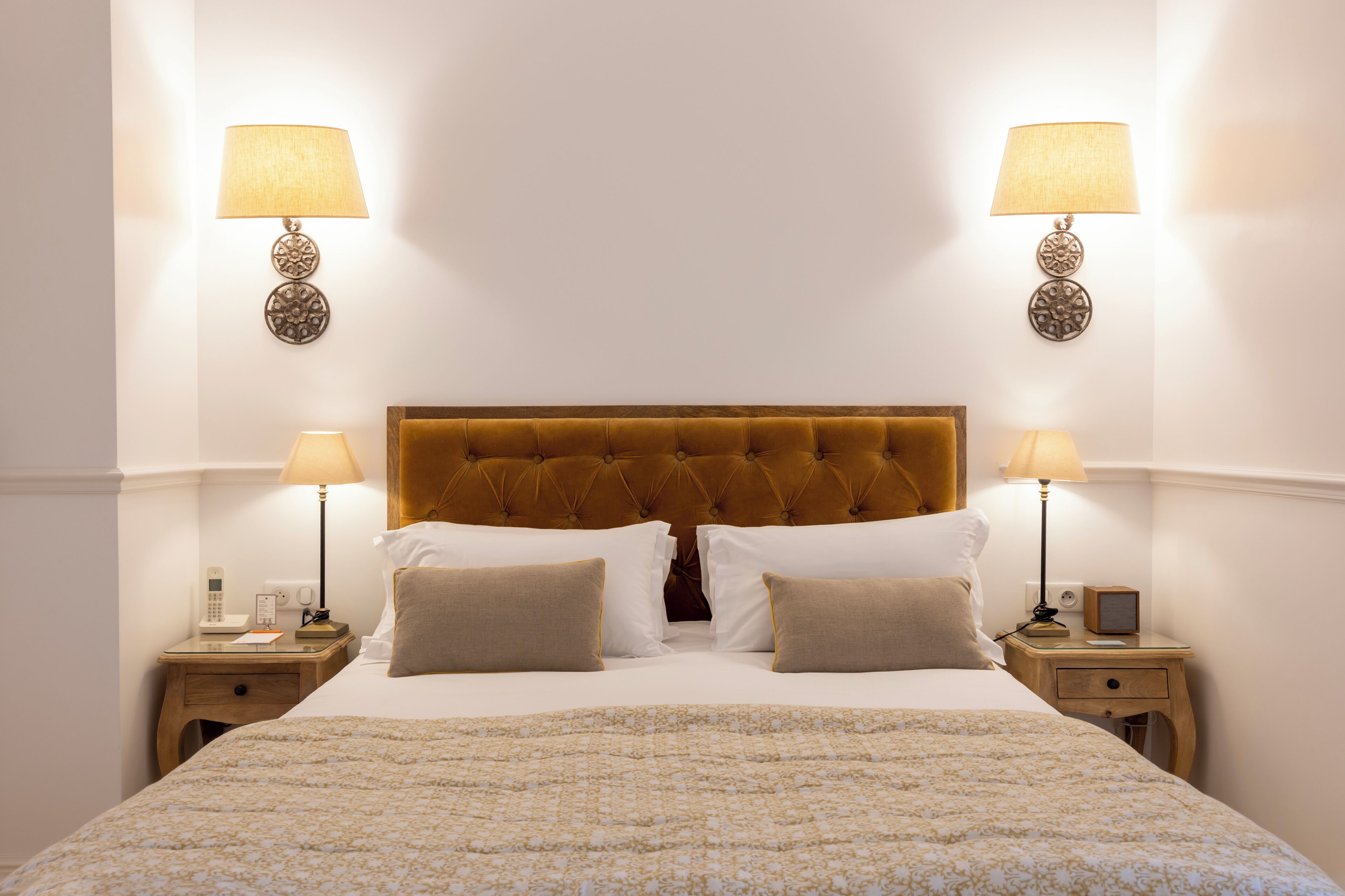 Un lit bien fait, recouvert d’une couvre-lit à motifs, avec deux lampes de chevet, un téléphone et une petite table de chaque côté.