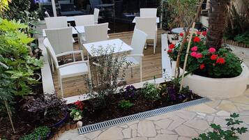 An outdoor seating area with wicker chairs and a table, surrounded by a variety of plants and flowers.