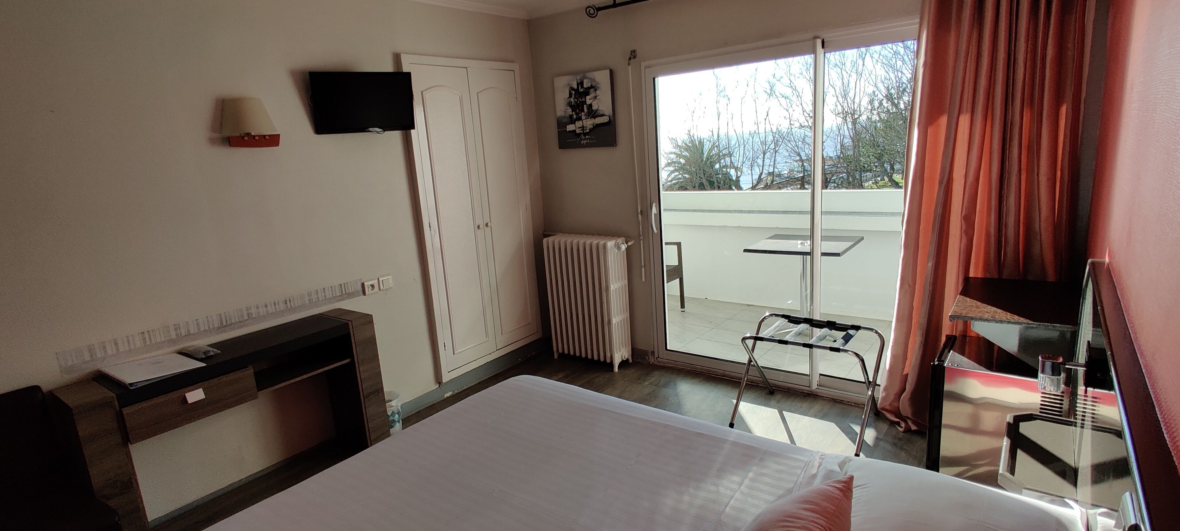 A hotel room with a bed, a desk, a chair, a radiator, a TV, and a sliding glass door leading to a balcony.
