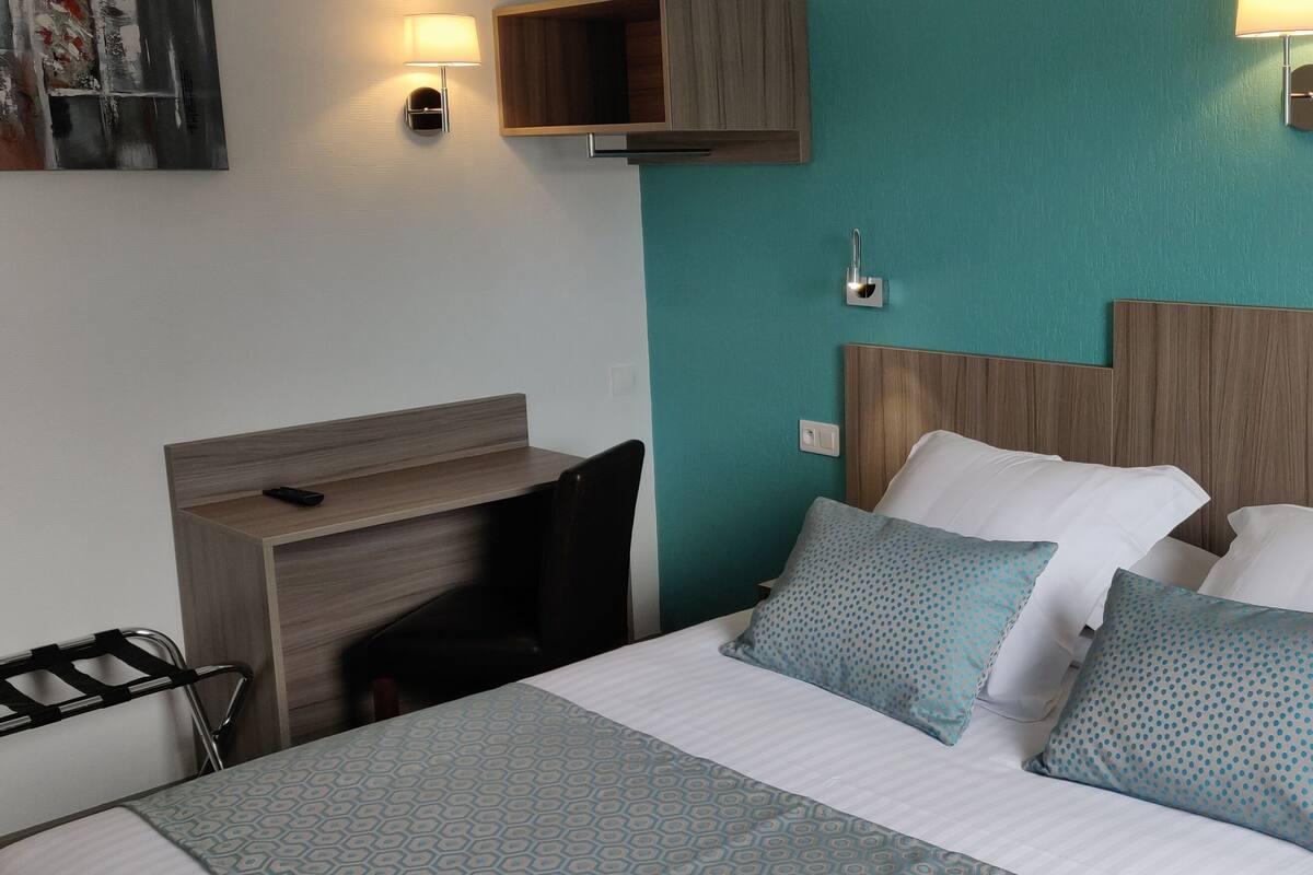 A hotel room with a bed, a desk, and a chair. There are two wall-mounted lamps and a painting on the wall.
