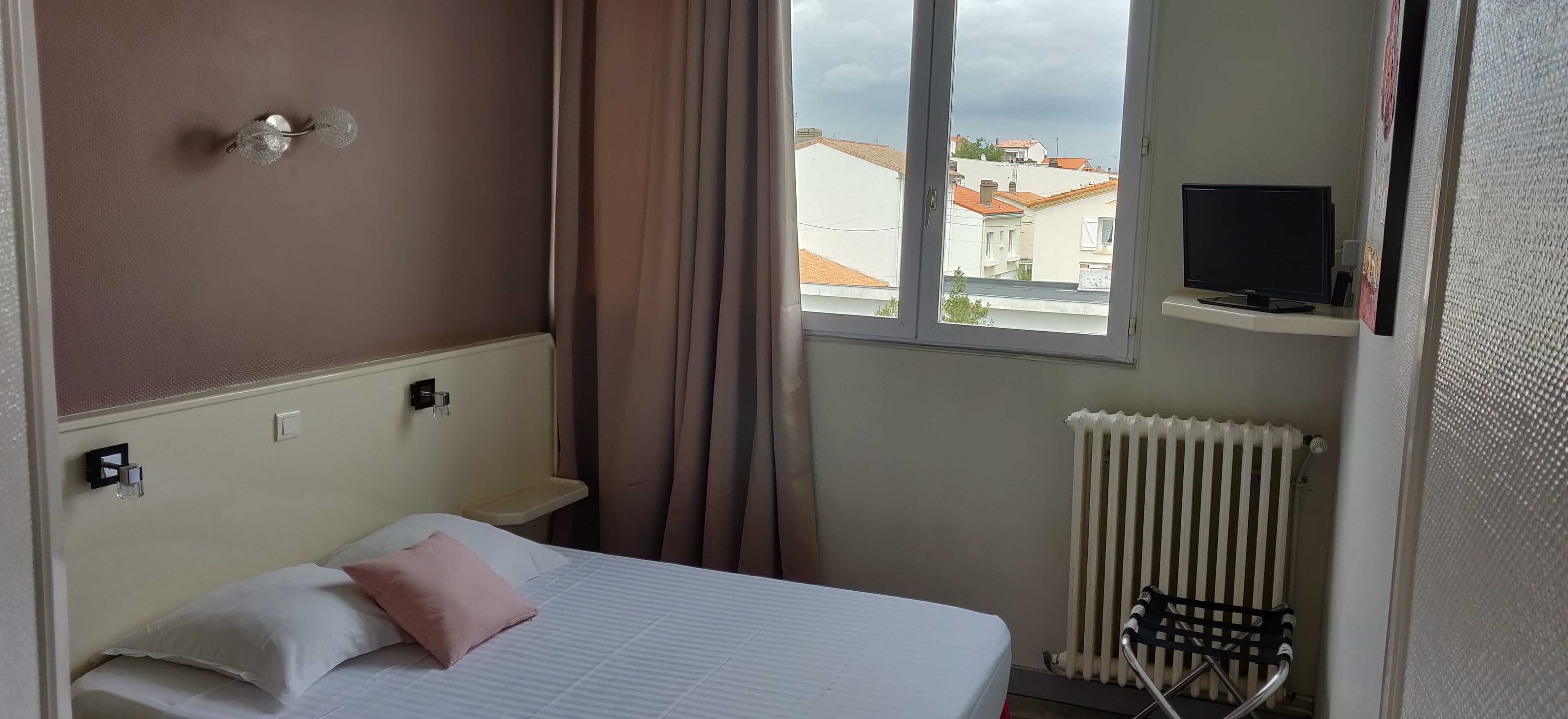 A hotel room with a bed, a window with a view of buildings, a TV on a stand, and a radiator.