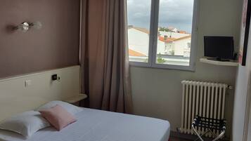 A hotel room with a bed, a window with a view of buildings, a TV on a stand, and a radiator.