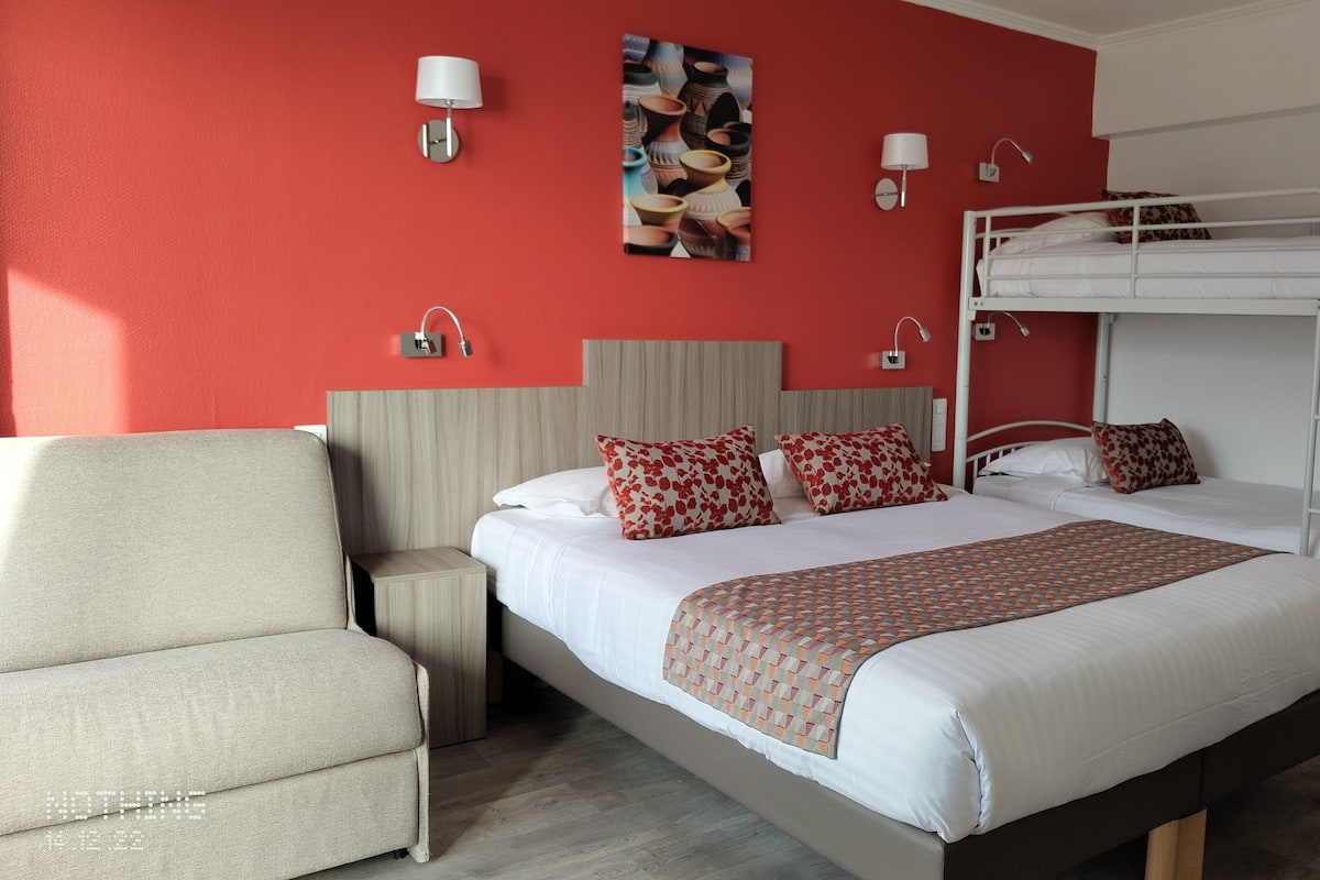 A hotel room with a bunk bed, a single bed, a sofa, a nightstand, and a wall-mounted lamp.