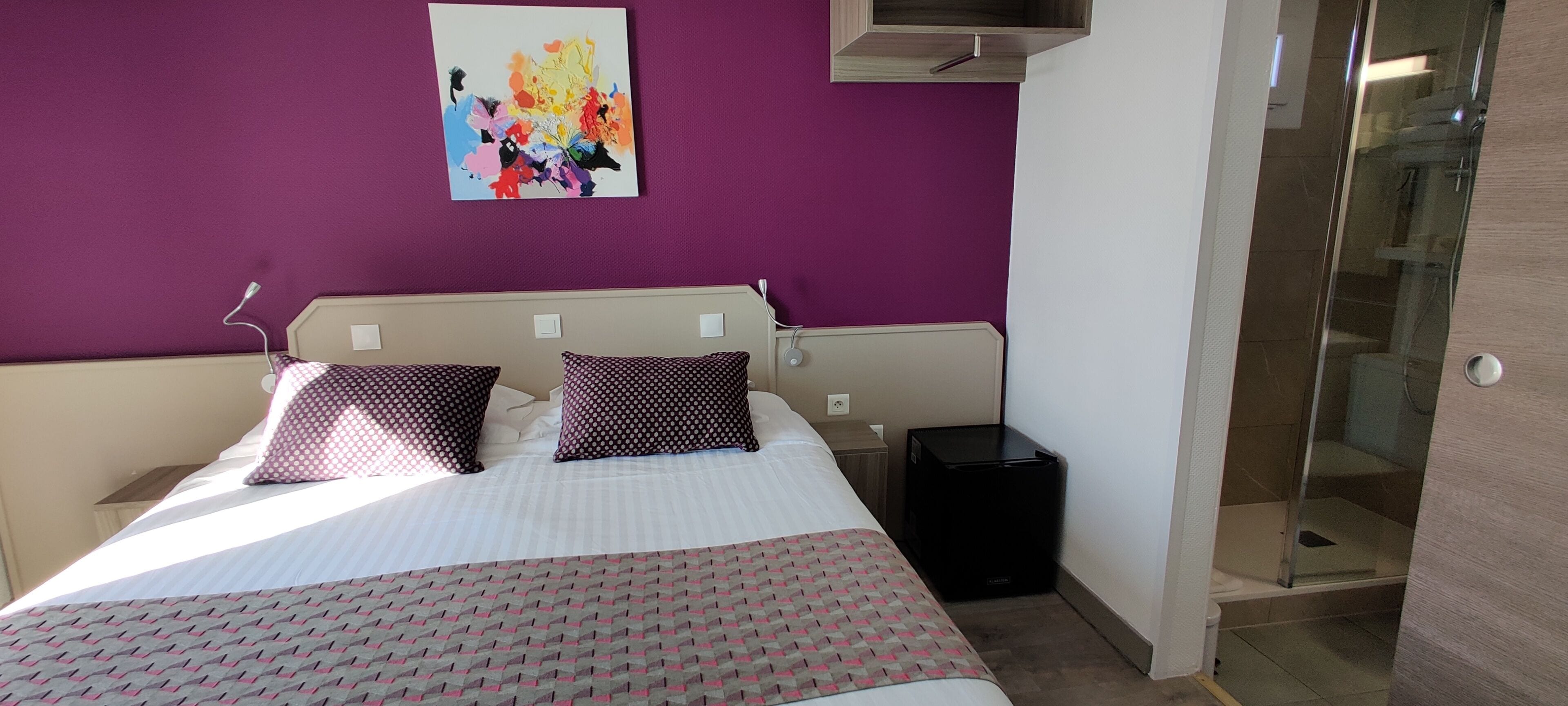 Une chambre à coucher avec un lit, deux oreillers, une table de chevet, une poubelle noire et un tableau abstrait aux couleurs vives accroché au mur.