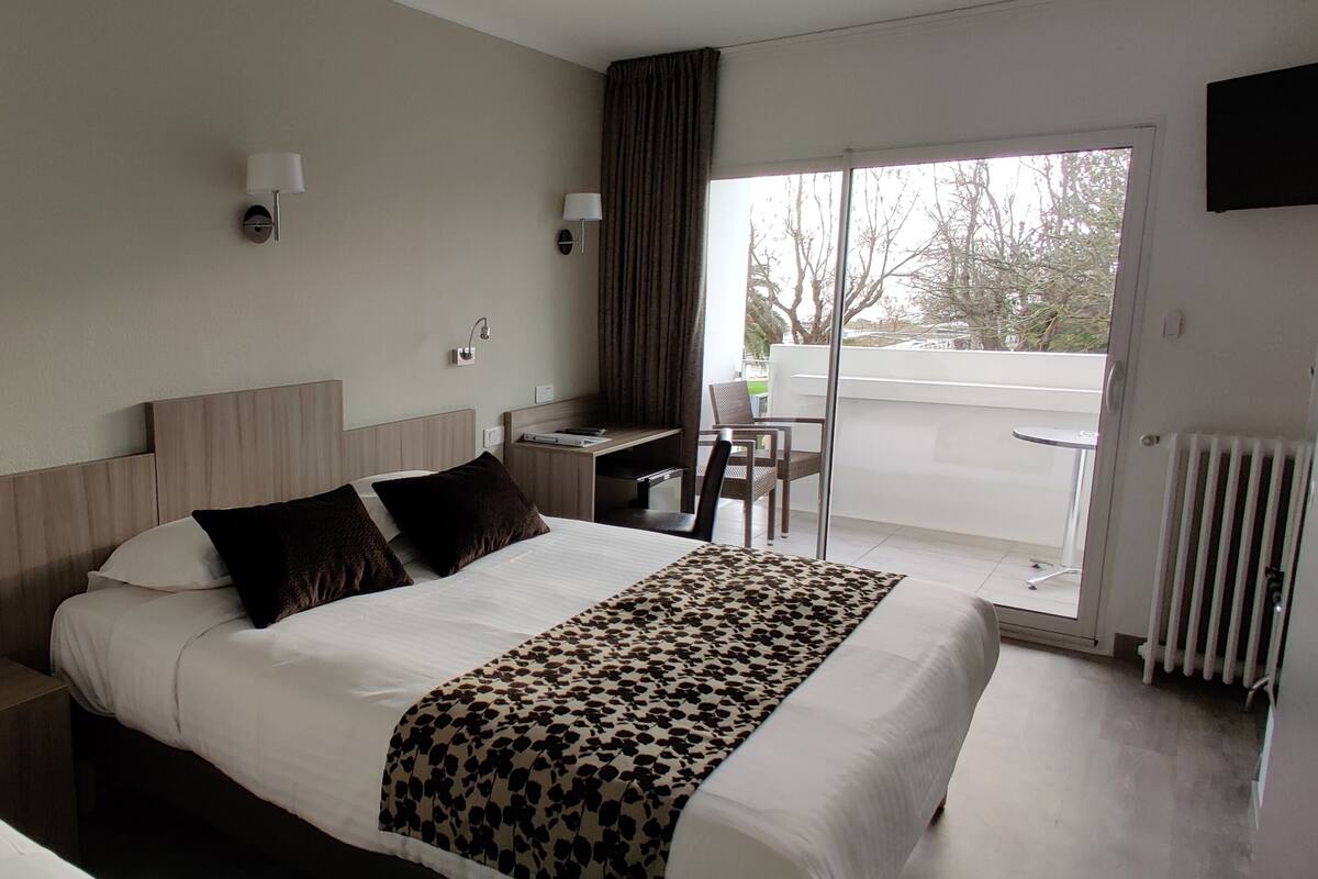 A hotel room with a bed, bedside tables, a desk, and a chair. There is a view of the outside through a glass door.