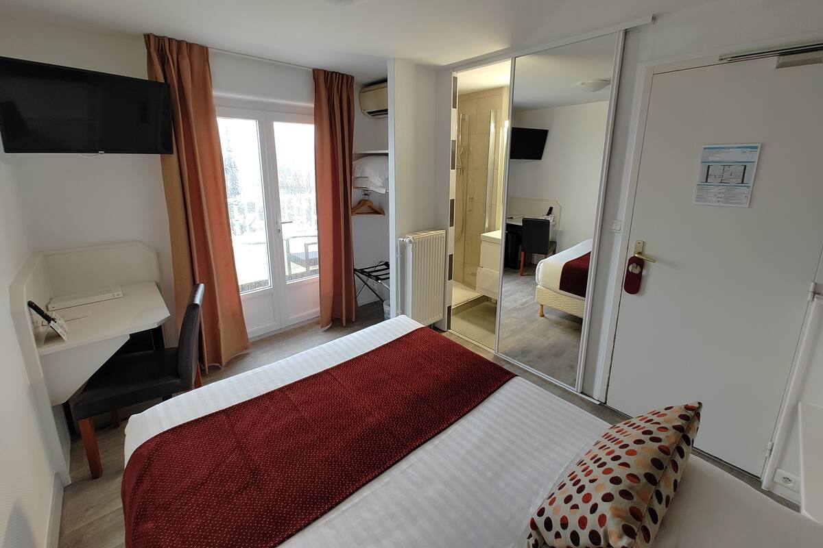 A hotel room with a bed, a desk, a chair, a TV, and a sliding door leading to a balcony.