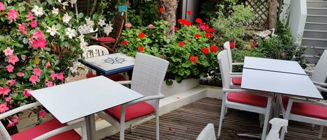 An outdoor patio with wicker furniture and red cushions, surrounded by lush greenery and vibrant flowers.
