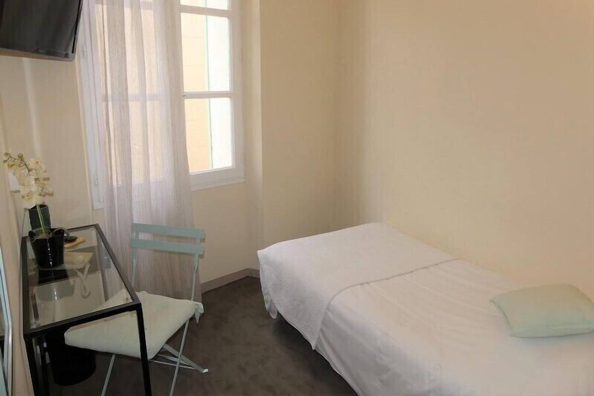 A small, clean room with a single bed, a desk with a chair, a TV mounted on the wall, and a window with sheer curtains.