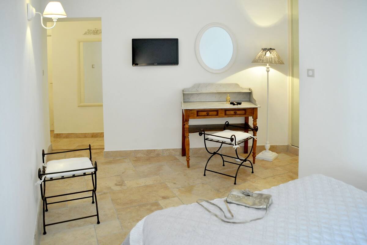 A hotel room with a bed, a bedside table with a lamp, a small desk with a mirror, a television, and a chair.