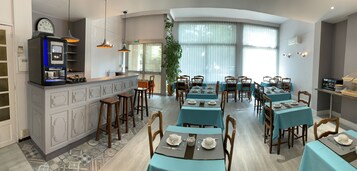 Un restaurant avec des nappes bleues, des chaises en bois et un coin bar agrémenté d’une horloge et d’une machine à café.