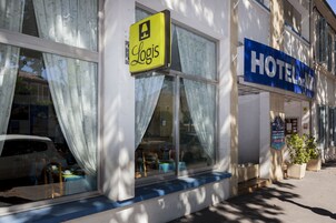 Un hôtel affichant les enseignes « Logis » et « HÔTEL », de grandes fenêtres avec des rideaux et un auvent bleu.