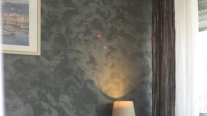 Una cama bien hecha con sábanas blancas y una sola almohada oscura, una mesita de noche con una lámpara y un cuadro en la pared.