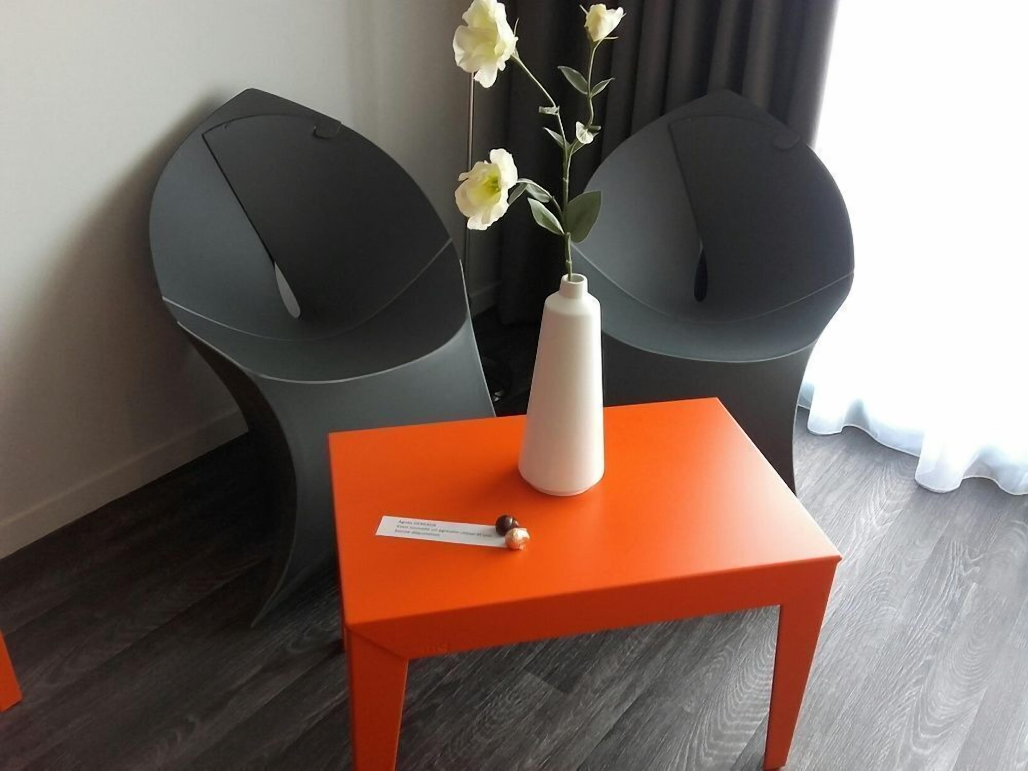 Een kamer met twee zwarte stoelen, een witte vaas met bloemen en een oranje tafel.