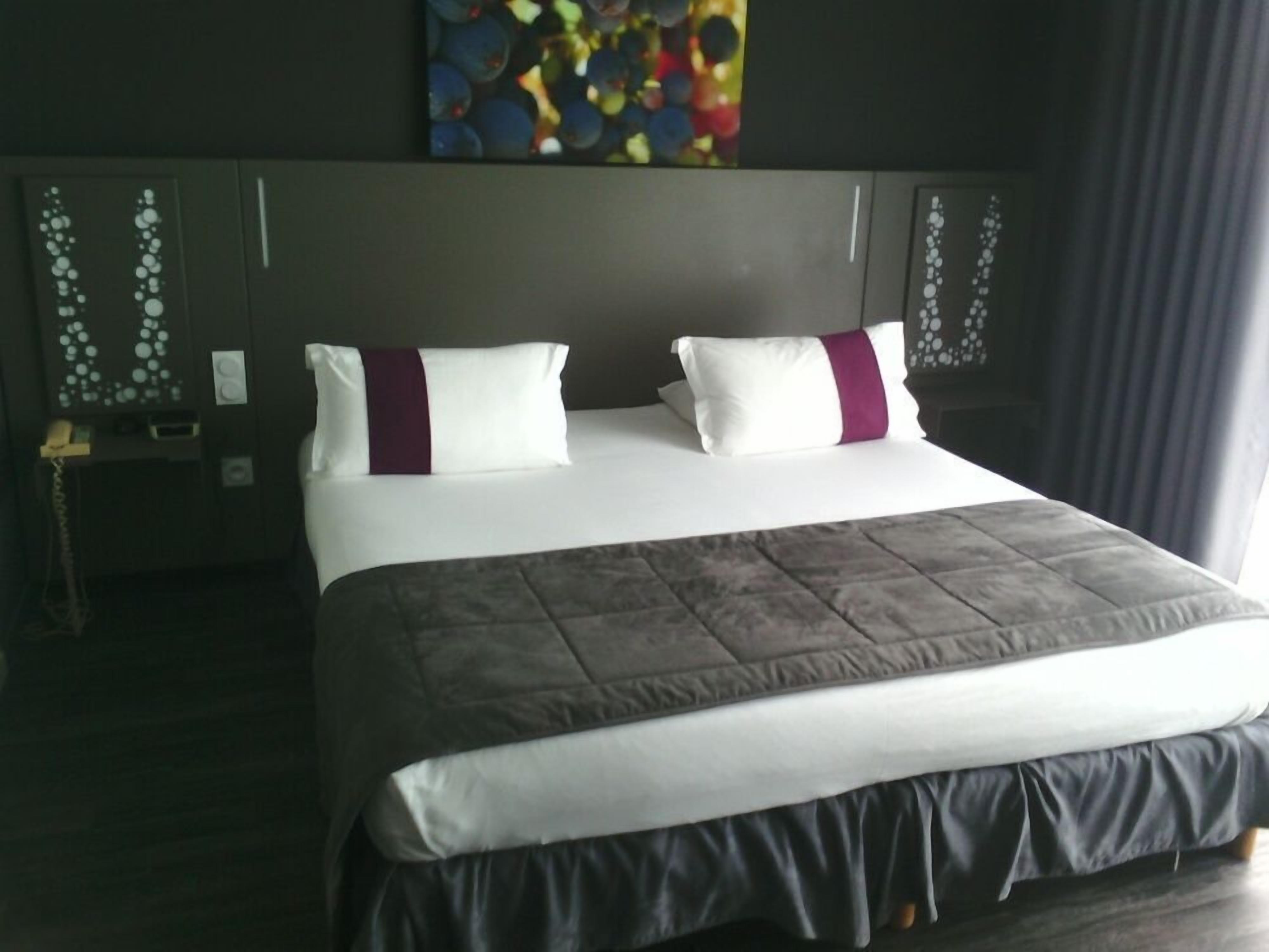 A hotel room with a large bed, a headboard with decorative elements, a bedside table with a lamp, and a painting on the wall.