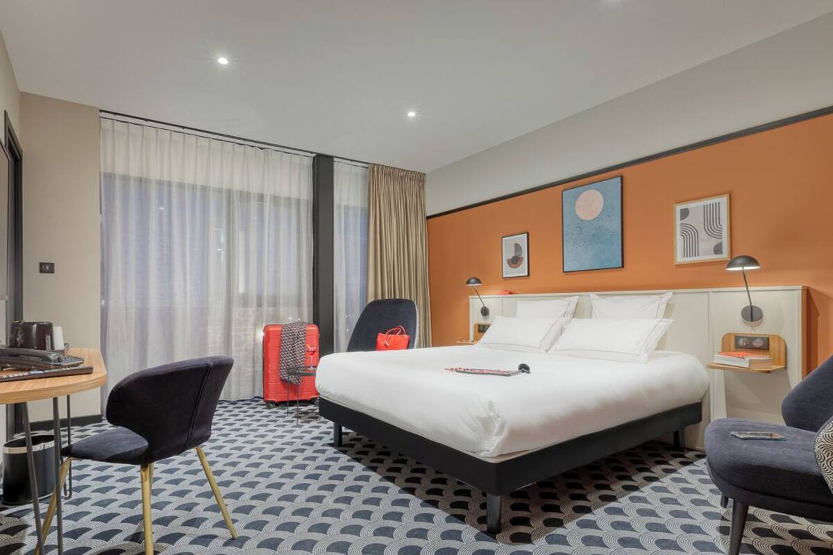 A hotel room with a bed, two chairs, a desk, a lamp, a radio, and a window with curtains.