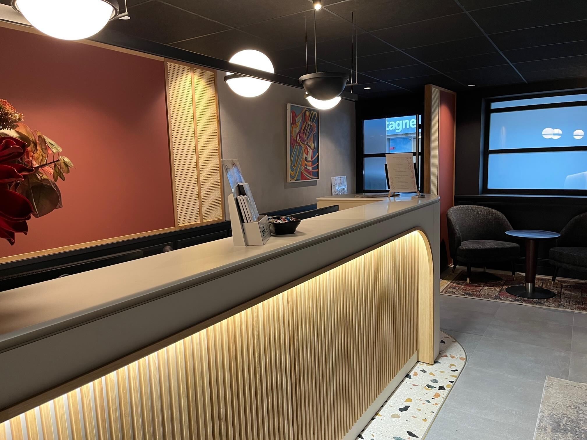 Zona de recepción con mostrador, área de descanso con dos sillones, planta decorativa y una pantalla grande mostrando un menú.