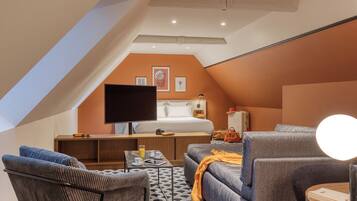 Ein modernes, kompaktes Zimmer mit schrägem Dach, einem Bett, einem Schreibtisch, einem Fernseher und Sitzgelegenheiten.