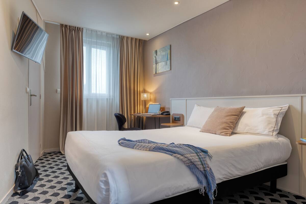 A hotel room with a large bed, a desk with a laptop, a chair, and a TV mounted on the wall.