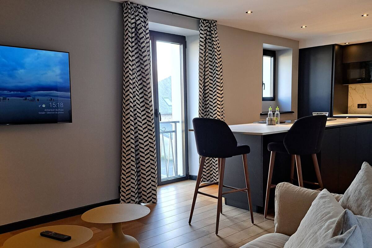 A modern hotel room with a sofa, two chairs, a small table, a TV mounted on the wall, and a kitchen area.