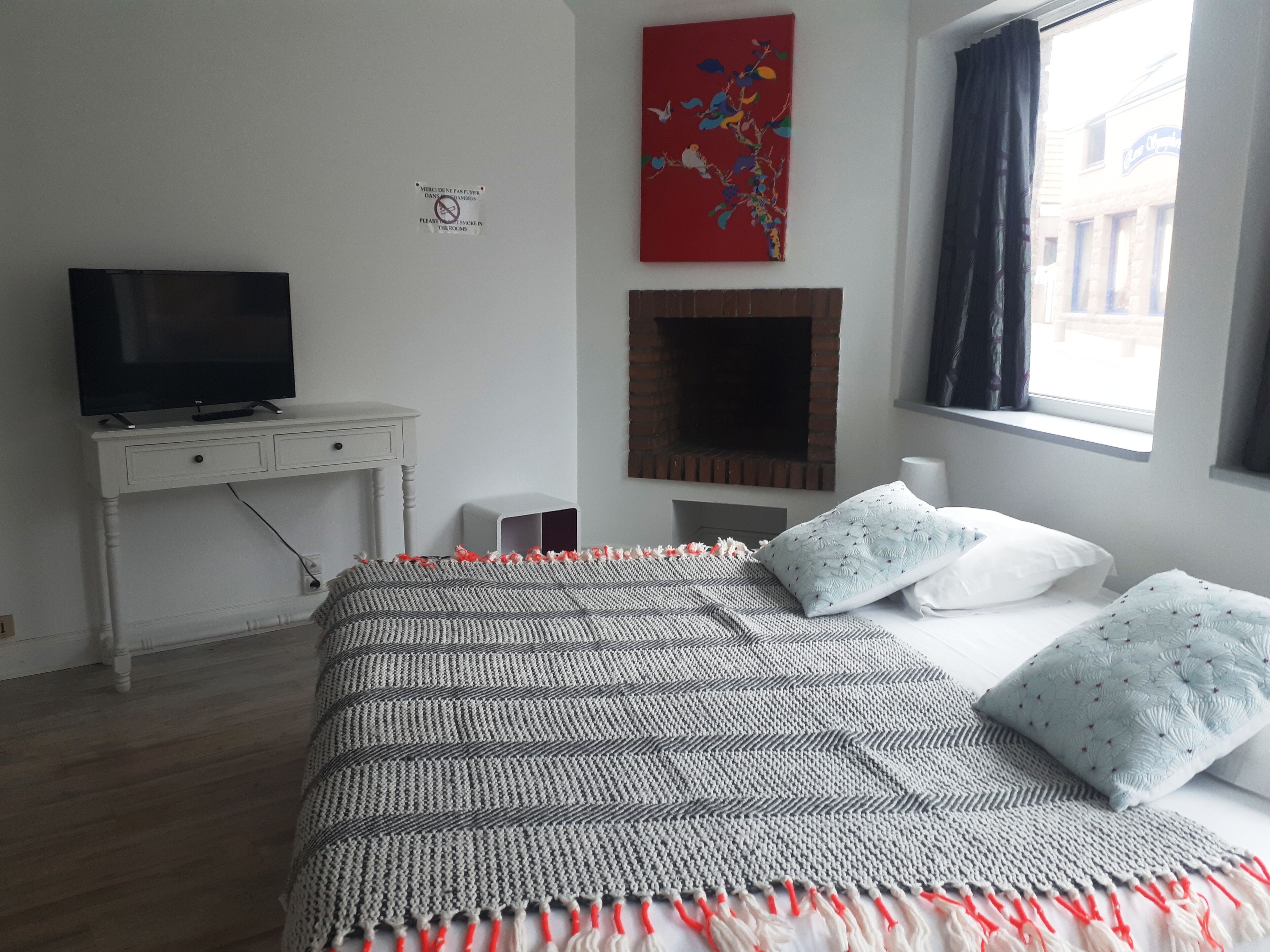 Une chambre à coucher avec un lit, un téléviseur posé sur une commode blanche, une cheminée et une fenêtre avec des rideaux.