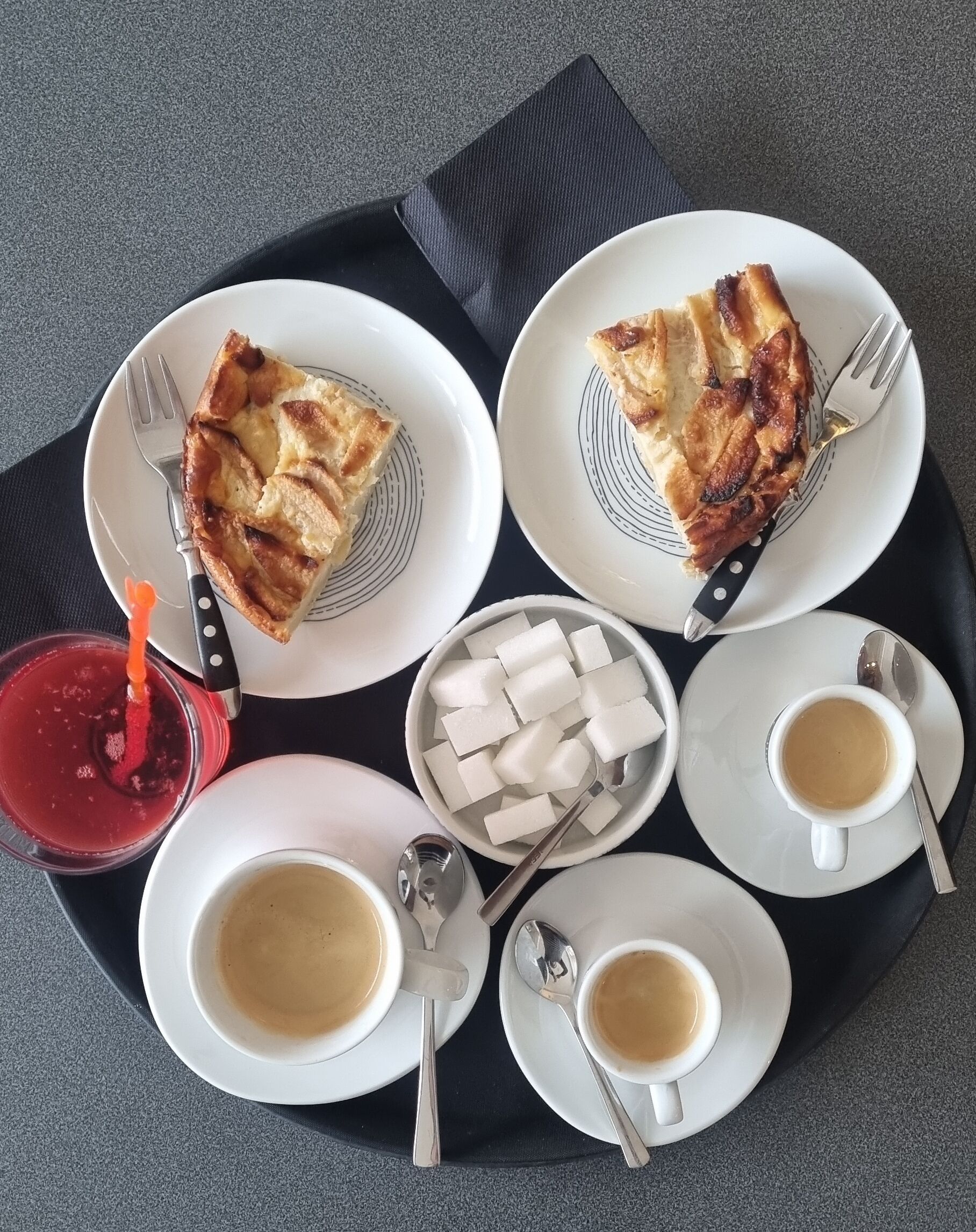 A tray with two plates of pie, a bowl of sugar cubes, two cups of coffee, and a glass of red drink.