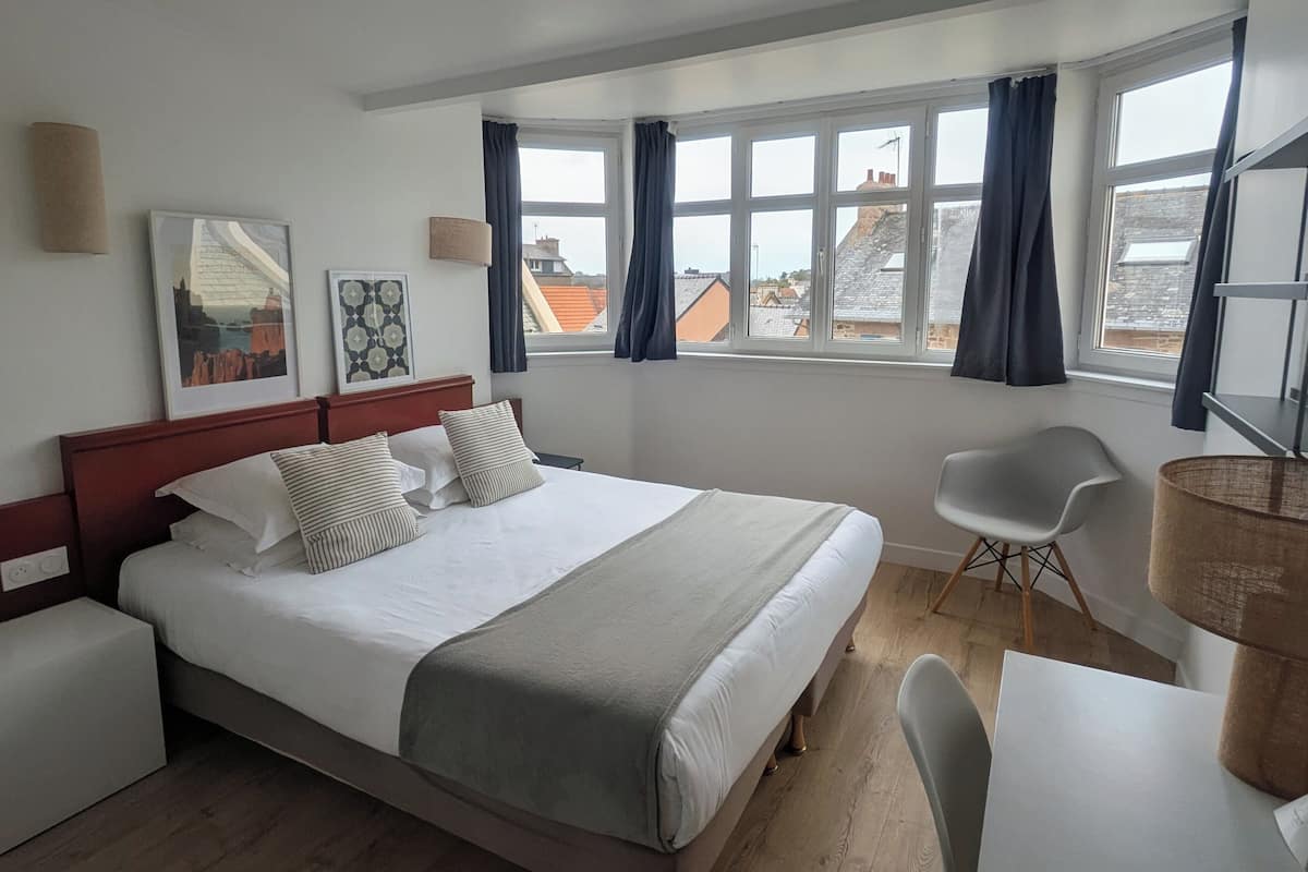 Ein Hotelzimmer mit Bett, Schreibtisch, Stuhl, Lampe und einem Fenster mit Vorhängen.
