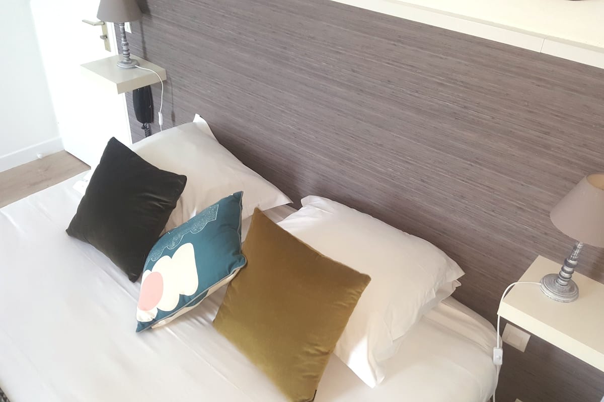 Ein ordentlich bezogenes Bett mit weißen Leinentüchern und drei Kissen in verschiedenen Farben.