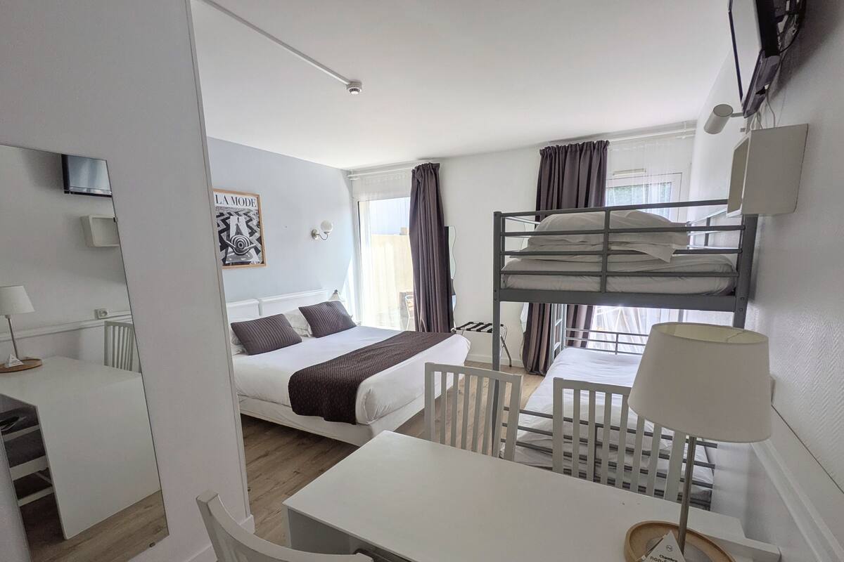 Ein kleines Hotelzimmer mit Etagenbett, einem Einzelbett, einem Schreibtisch mit Stuhl, einem kleinen Tisch mit Lampe und einem Fenster mit Vorhängen.