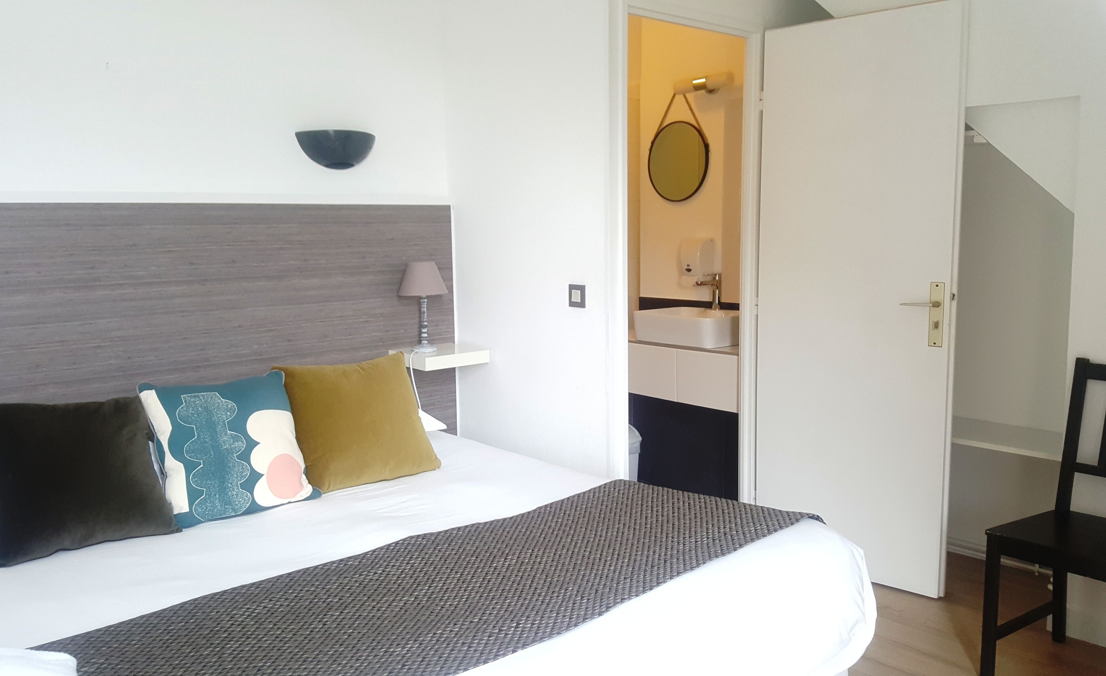 Habitación de hotel con cama, mesita de noche con lámpara, luz empotrada en la pared y puerta que da a un baño.