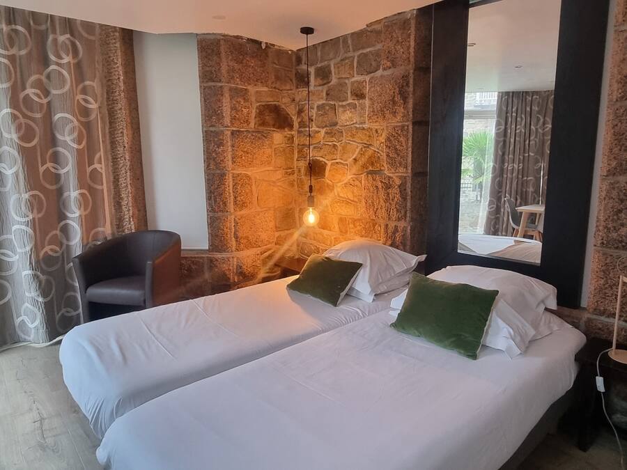 A hotel room with two beds, a stone wall, a chair, a table, and a lamp.