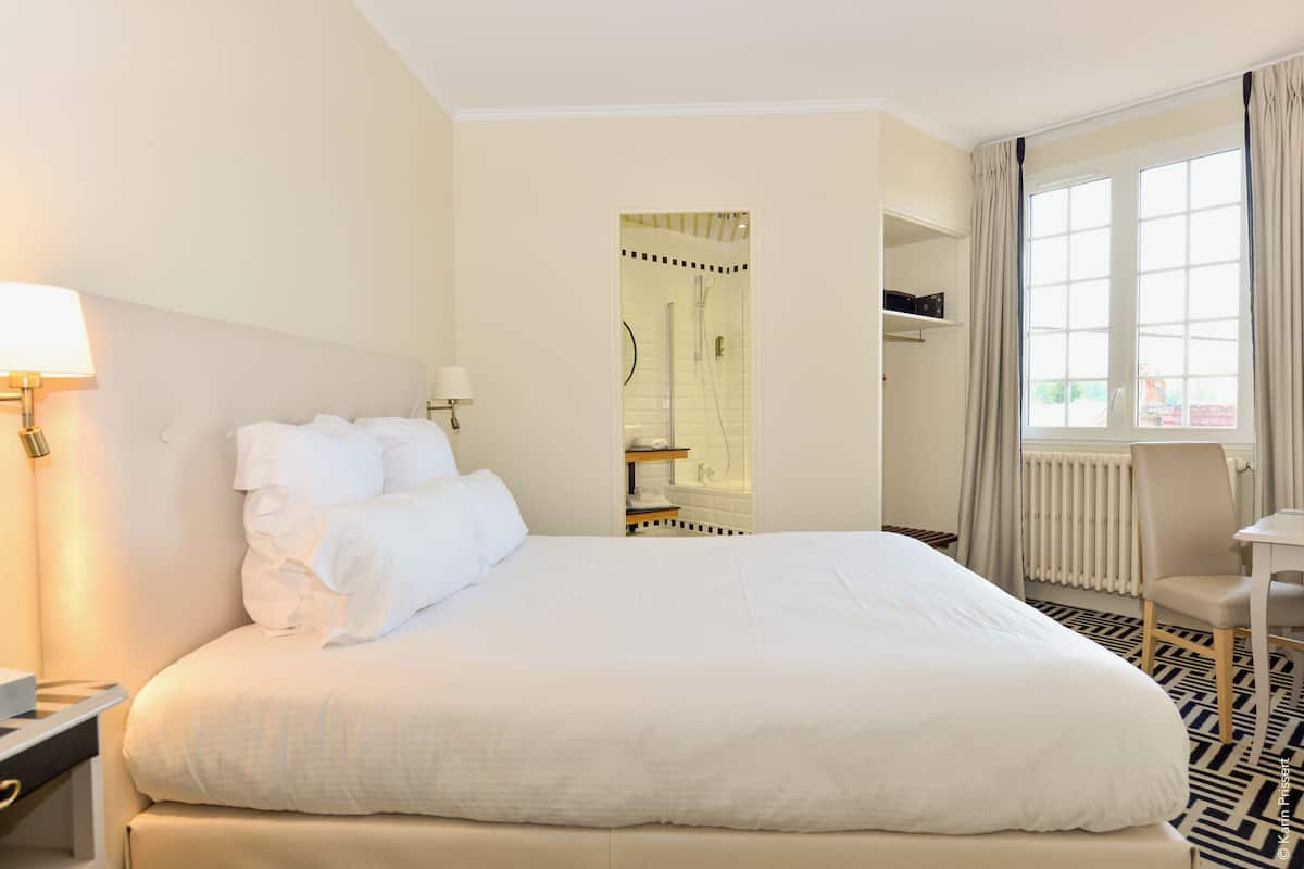 A hotel room with a large bed, a bedside table, a lamp, a window with curtains, and a bathroom visible through an open door.