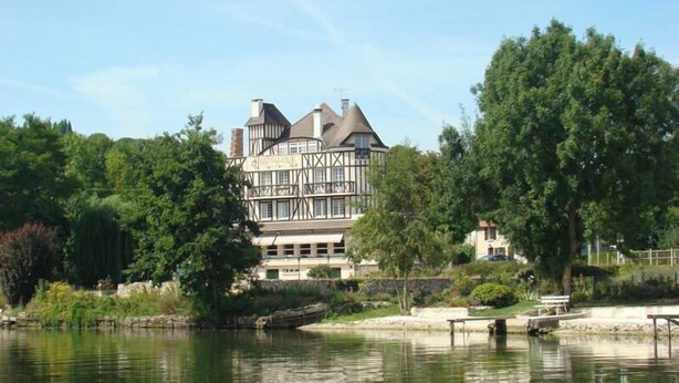 Un edificio grande e histórico con una torre destacada y numerosas ventanas, ubicado junto a un lago tranquilo con un muelle y bancos.