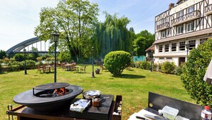 Un patio trasero con una parrilla, mesas y sillas, rodeado de follaje y un edificio con vigas de madera a la vista.