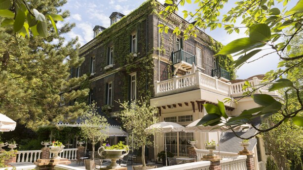 A two-story building with a balcony, outdoor seating area with umbrellas, and a garden with potted plants.