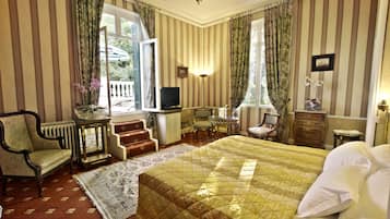 Quarto de hotel com uma cama grande, televisão, escrivaninha com cadeira, mesa pequena com um vaso de flores e janela com cortinas.