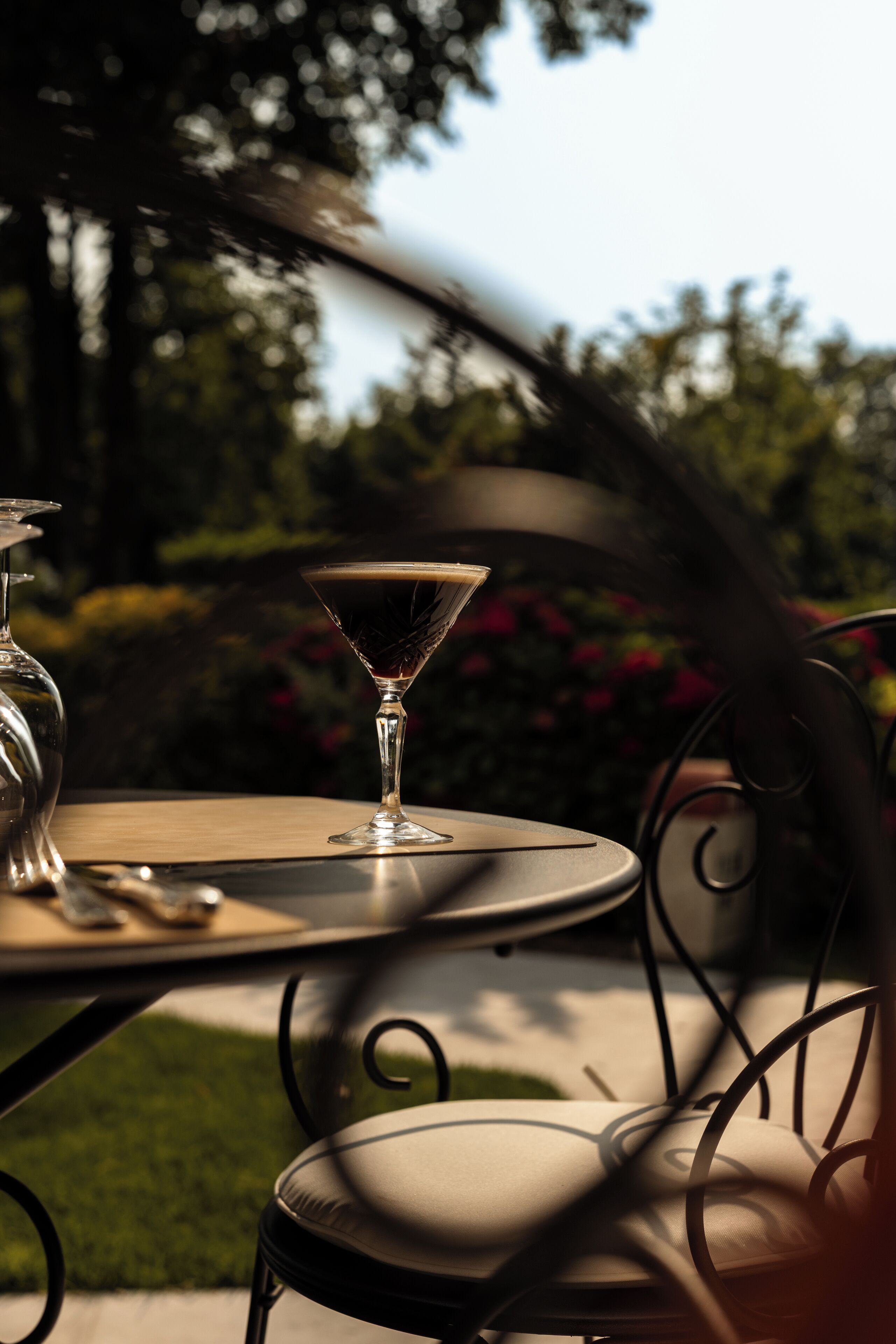 Une table avec un verre à martini et une bouteille, disposée à l’extérieur, avec un jardin en arrière-plan.