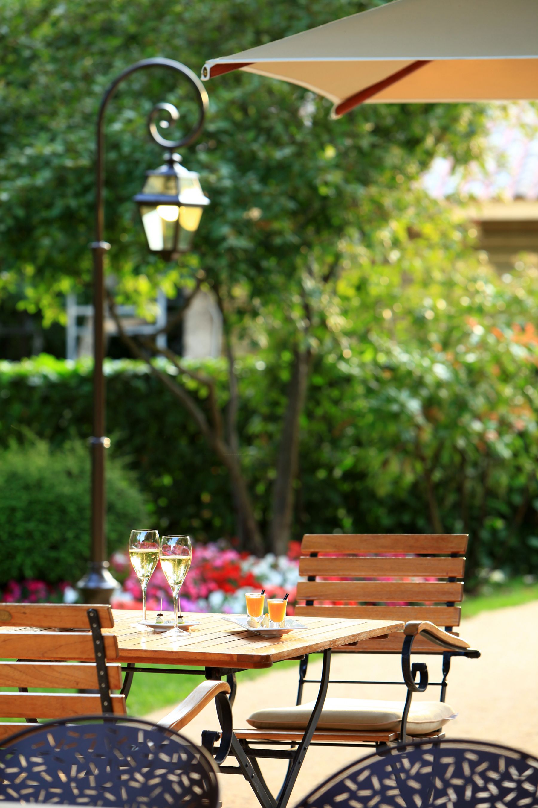 Une terrasse extérieure avec une table et des chaises en bois, une lampe en métal et un verre de jus d’orange.