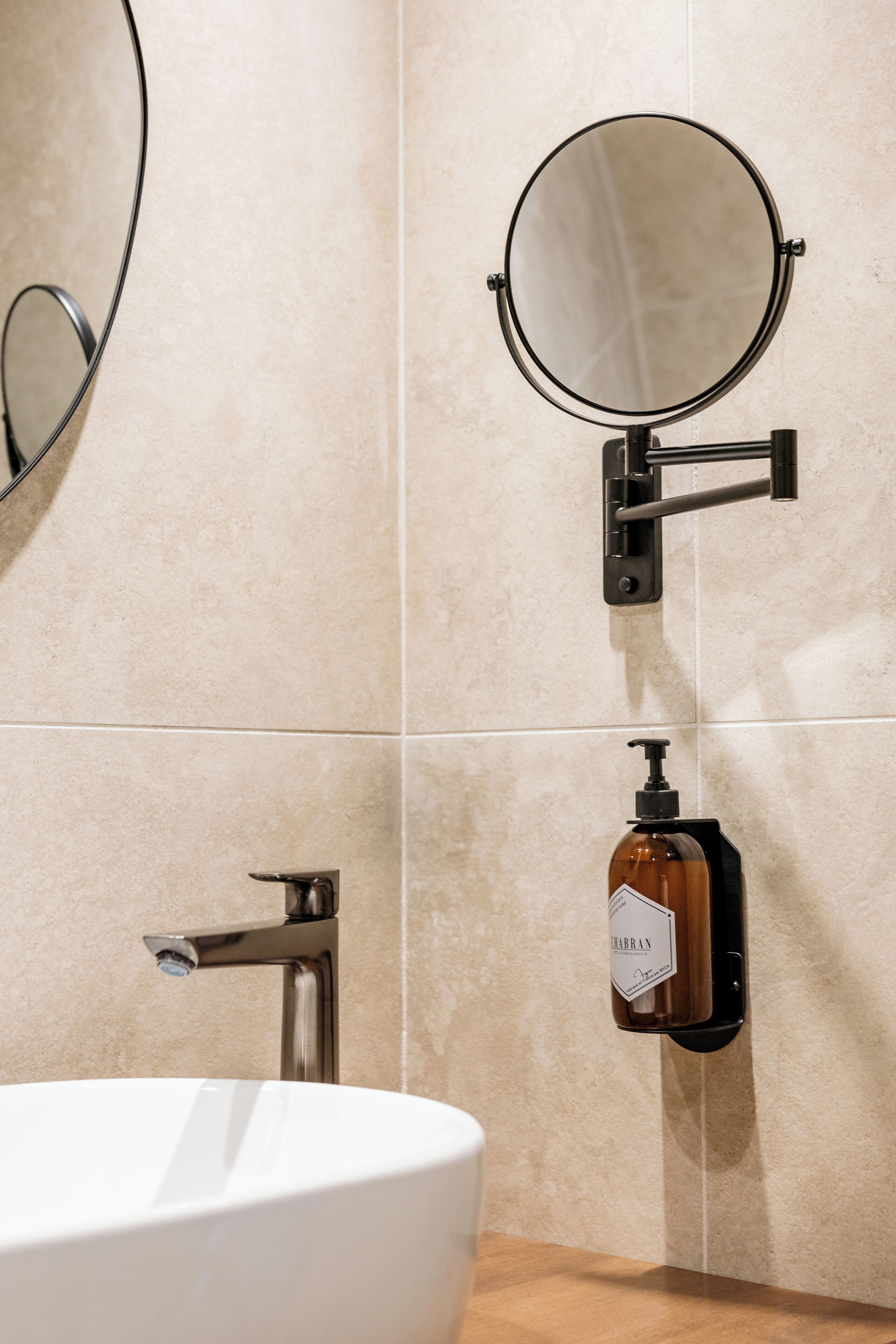 A bathroom with a white sink, a round mirror, and a soap dispenser mounted on the wall.